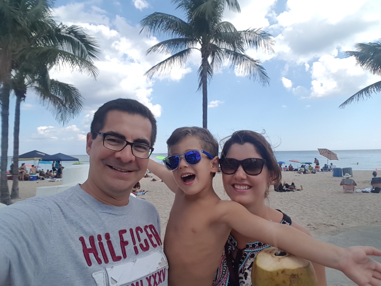 José Eduardo, seu filho de 4 anos e a esposa Danielle estão em uma selfie cujo fundo é uma praia. O céu está azul e Danielle segura uma água de coco. O filho do casal está no colo de José e posa para foto de braços abertos. 