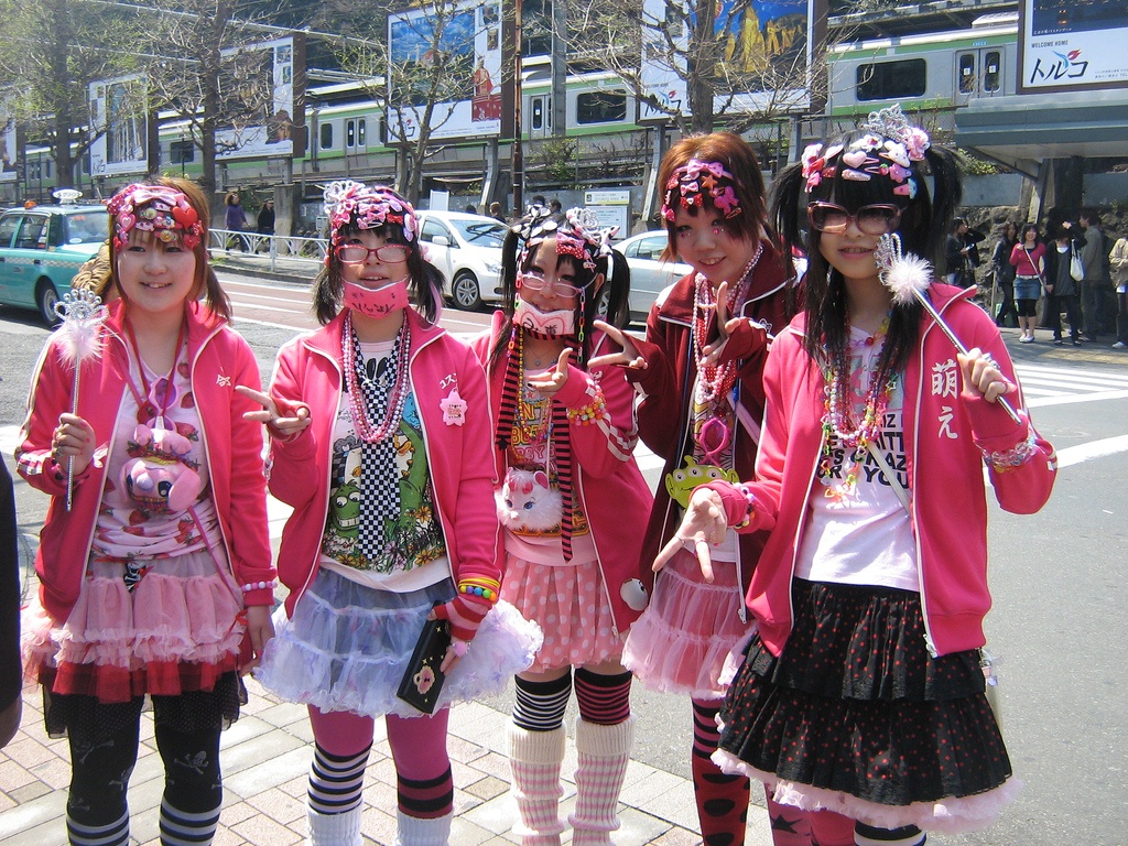 Um grupo de cinco meninas japonesas vestidas no estilo Dekora, com sobreposições e roupas rosas, sorriem para a foto.