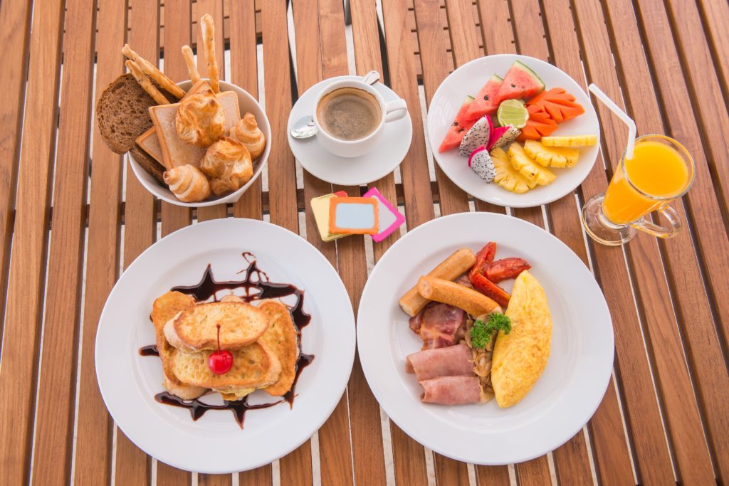 Uma xícara de café, um prato com panquecas, um prato com frutas e um copo de suco de laranja sobre mesa de madeira compõe um café da manhã