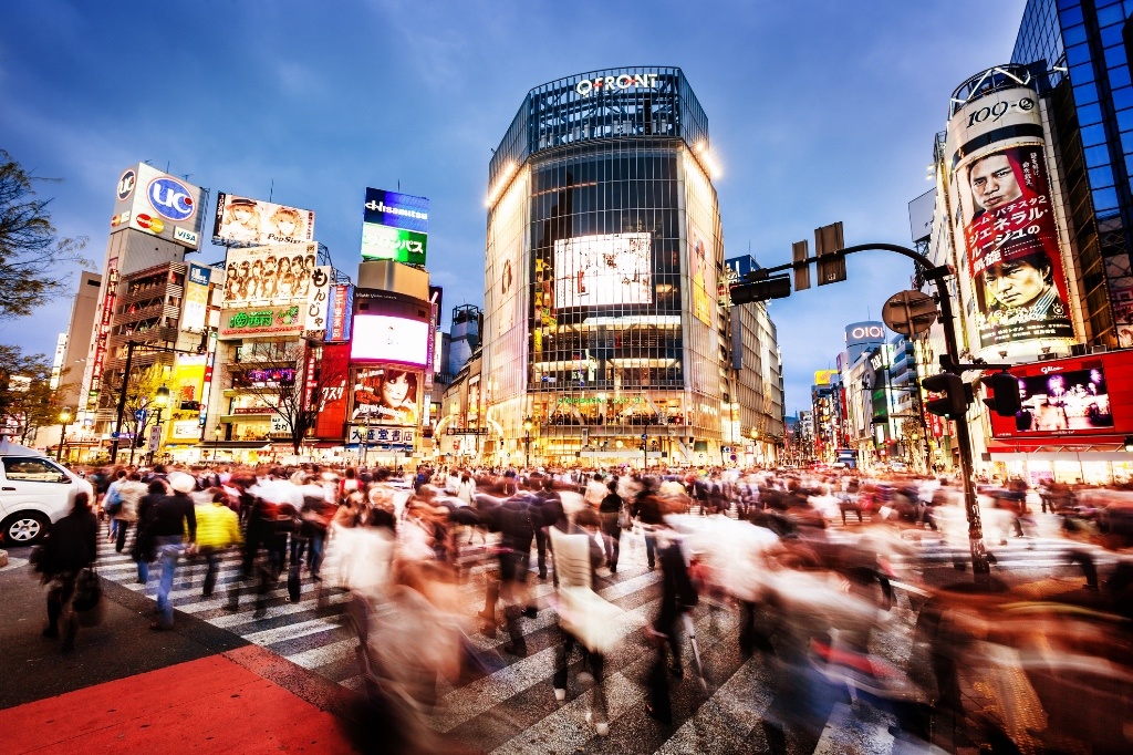 Imagem desfocada de Tóquio na qual aparece uma esquina com muitas pessoas passando e prédios imponentes. A imagem tem uma poluição visual contundente, mas mesmo assim é muito bonita. 