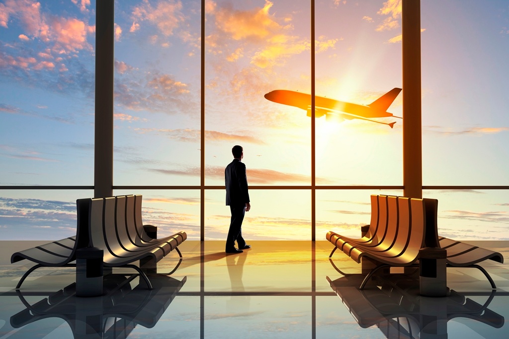 Imagem de um jovem parado no saguão de um aeroporto observando um avião decolar. Ao fundo o por do sol dá um ar bucólico e esperançoso à imagem.